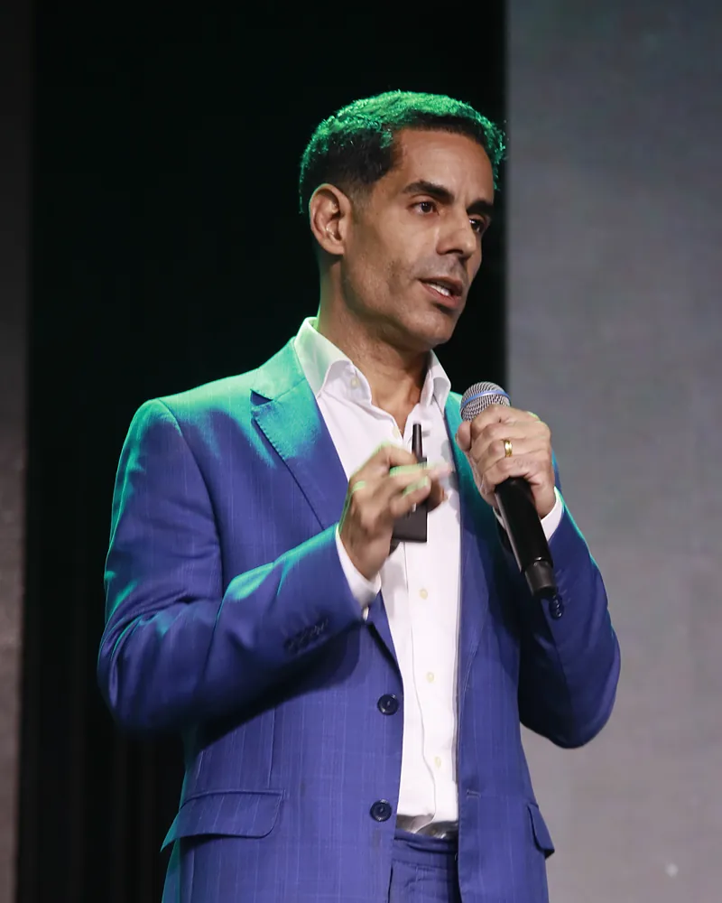 Dr. Bruno Menezes palestrando em congresso médico, de terno azul e microfone na mão